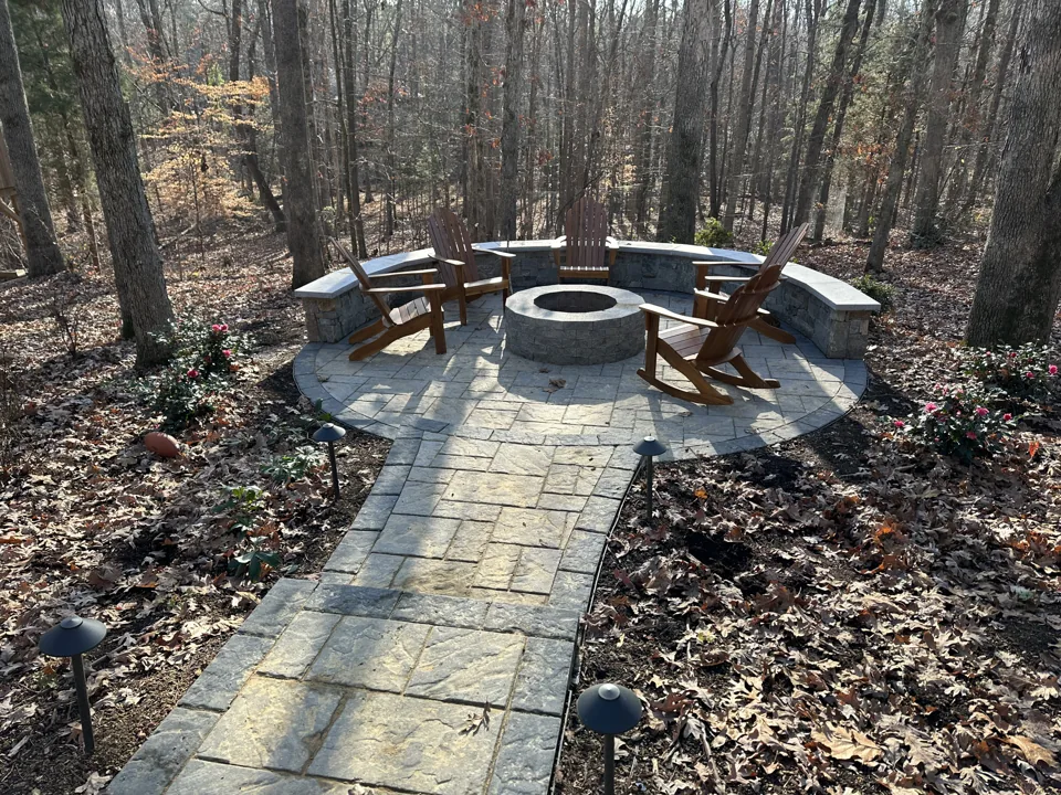 A paver walkway leading to a circular fire pit and Adirondack chairs among trees
